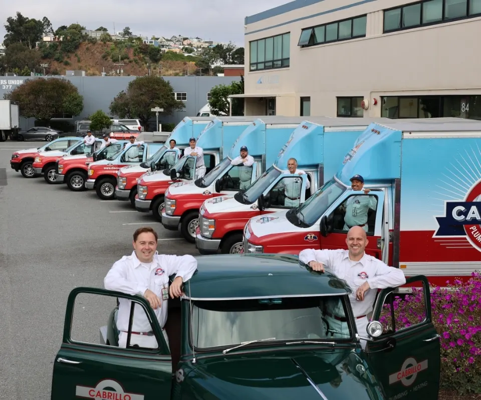 The real Cabrillo Plumbing, Heating & Air crew at the San Francisco shop — two technicians on the restored vintage green pickup with the rest of the team posted beside the red-and-white service fleet.